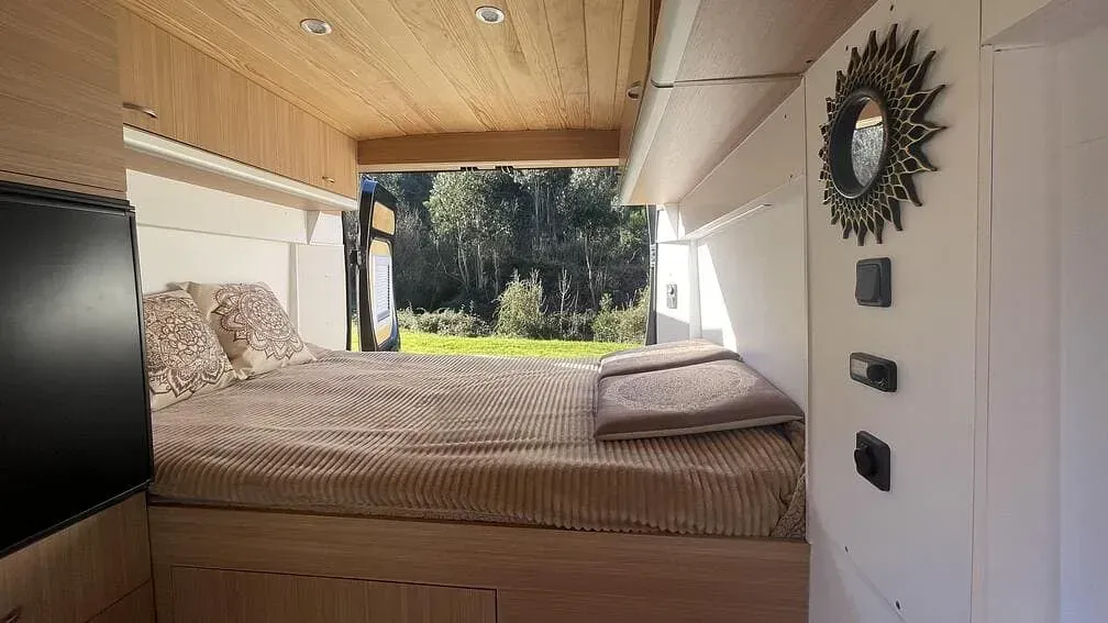 Interior de una autocaravana con cama, ventana con vistas al verde paisaje y paneles de madera. Un espejo con efecto rayos de sol cuelga de la pared.