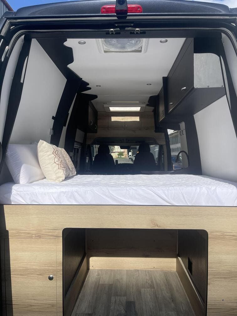 Interior de una furgoneta reformada con cama, armarios y claraboya. Detalles en negro, blanco y madera clara.