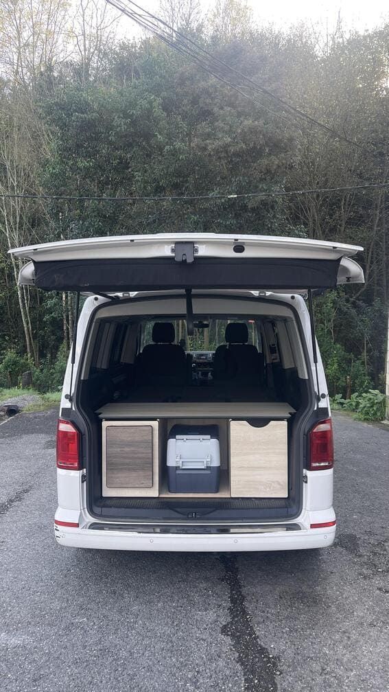Una camioneta blanca con el maletero abierto está estacionada al costado de la carretera.