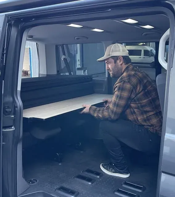 Hombre instalando una cama plegable en una furgoneta, probablemente para acampar. Lleva camisa a cuadros, sombrero y pantalones negros. La furgoneta es gris oscuro.
