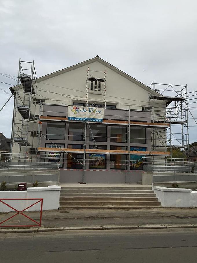 Bâtiment avec échafaudage en cours de rénovation ; façade grise, marches, ciel nuageux.