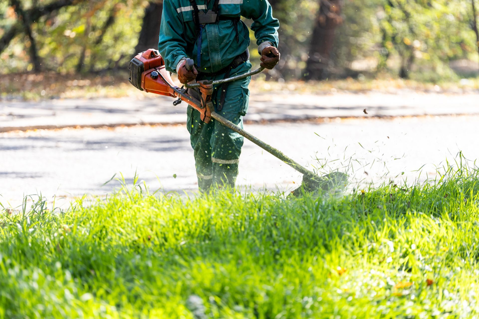 Une personne en salopette verte utilise une débroussailleuse pour tailler les hautes herbes près d'un chemin.