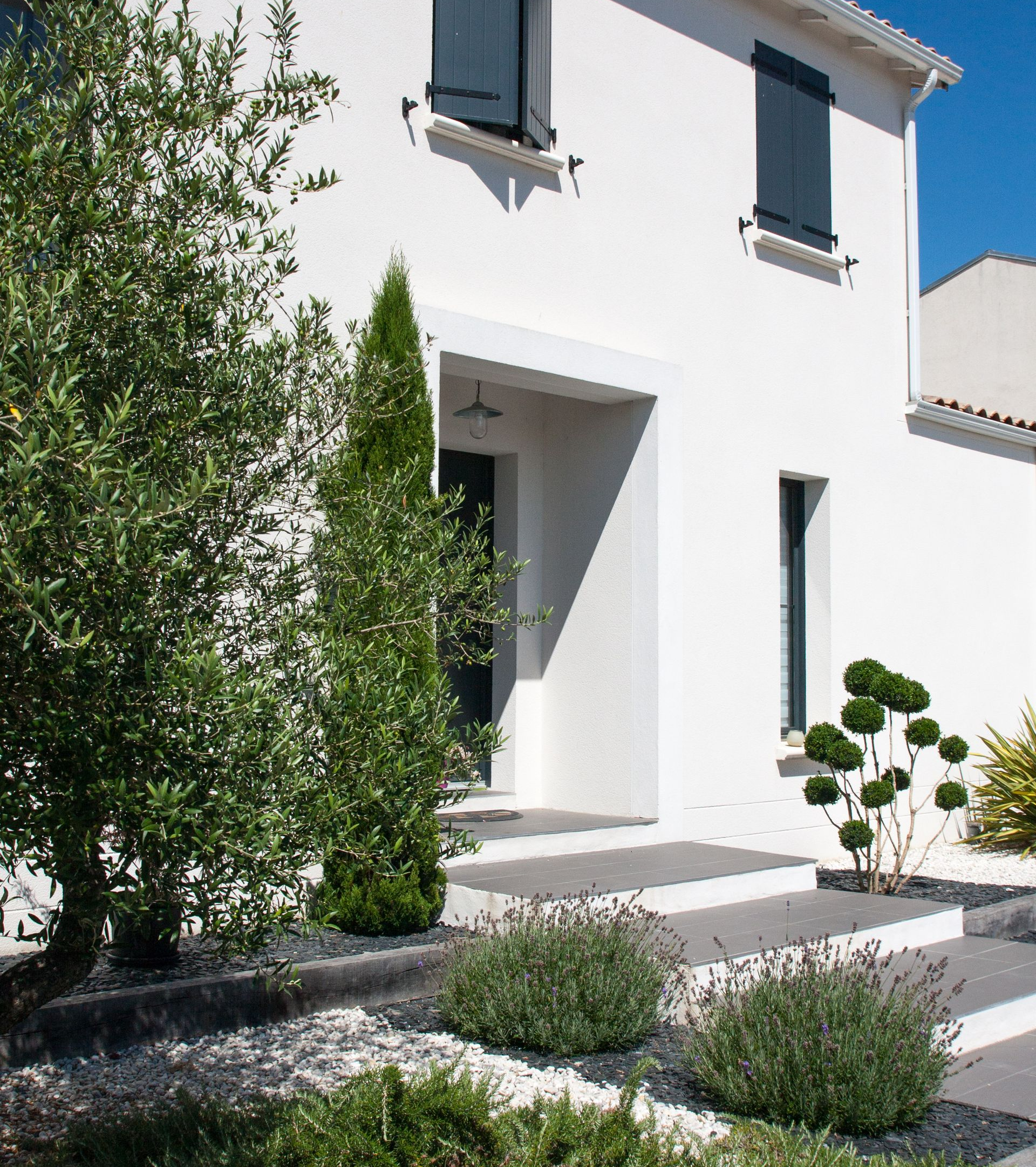 Maison en stuc blanc avec volets foncés, marches d'entrée et jardin paysager.
