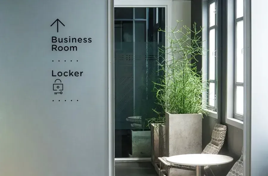 Couloir avec un panneau indiquant la salle de réunion et des casiers, une plante en pot, une chaise et une petite table ronde près des fenêtres.