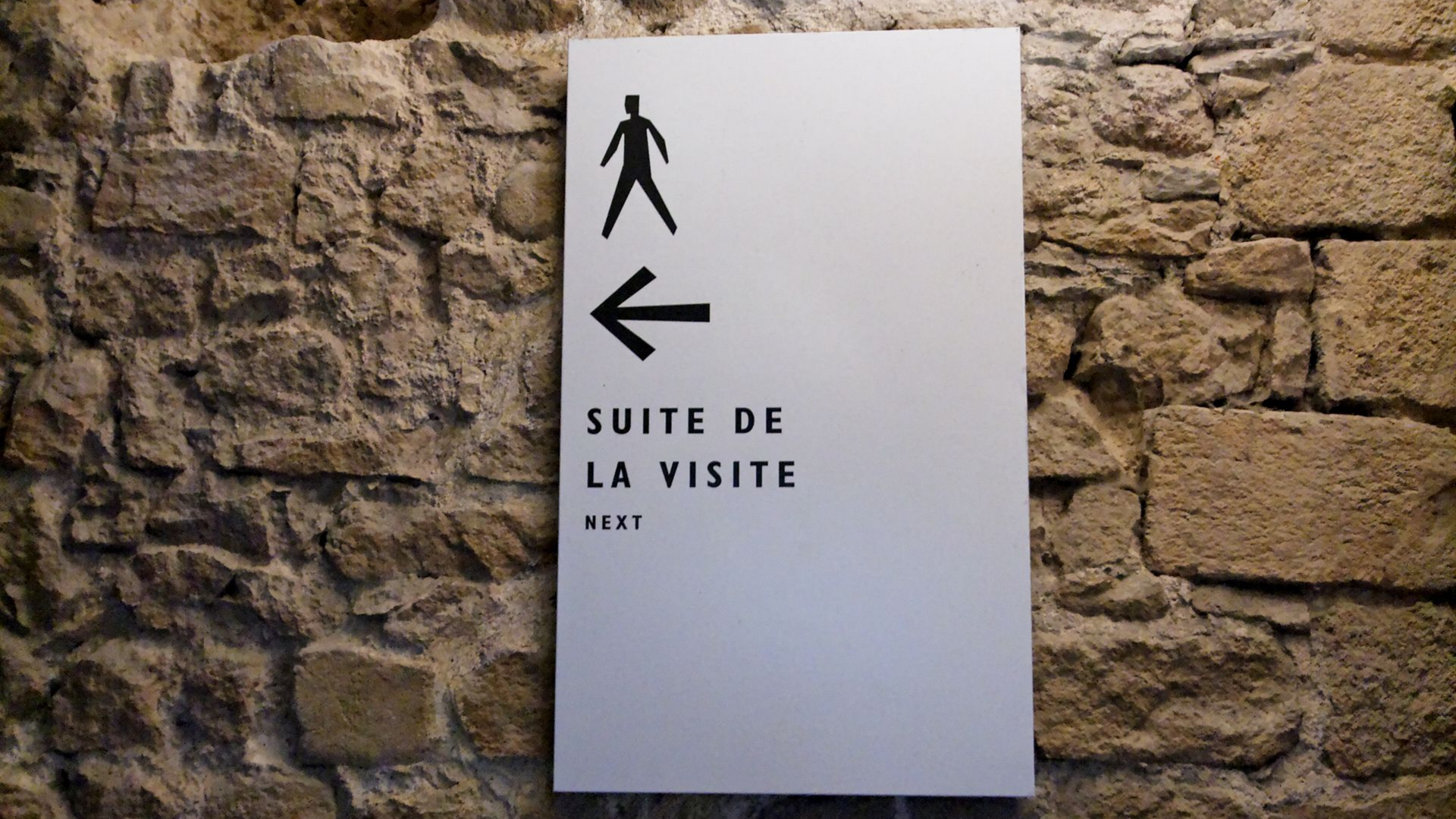 Panneau blanc sur un mur en pierre avec un piéton, une flèche vers la gauche et le texte français « SUITE DE LA VISITE. »