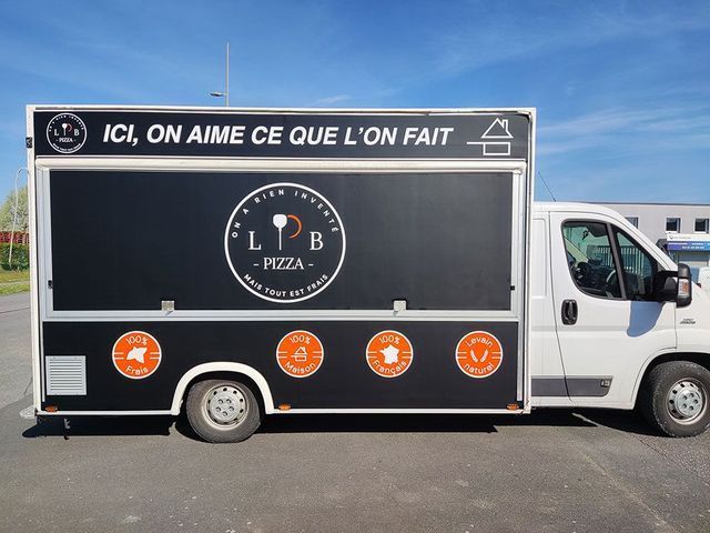 Food truck blanc avec un flanc noir façon tableau noir et du texte en français, garé en plein air sur un parking.