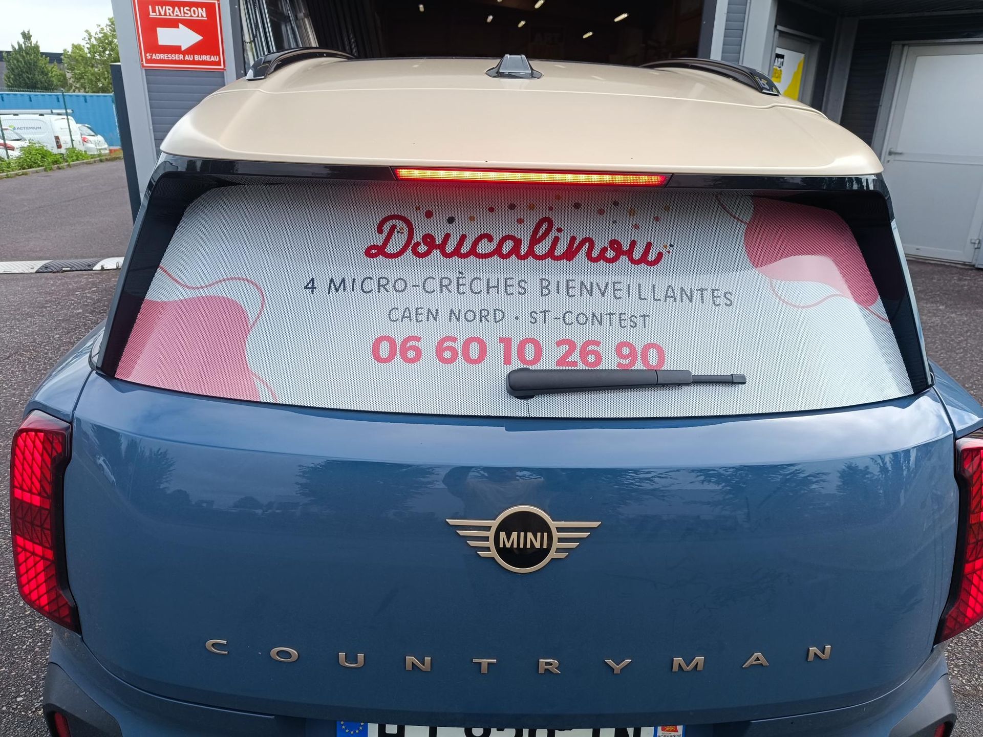 Arrière d’une Mini Countryman bleue avec une pancarte sur la vitre et un numéro de téléphone affiché.
