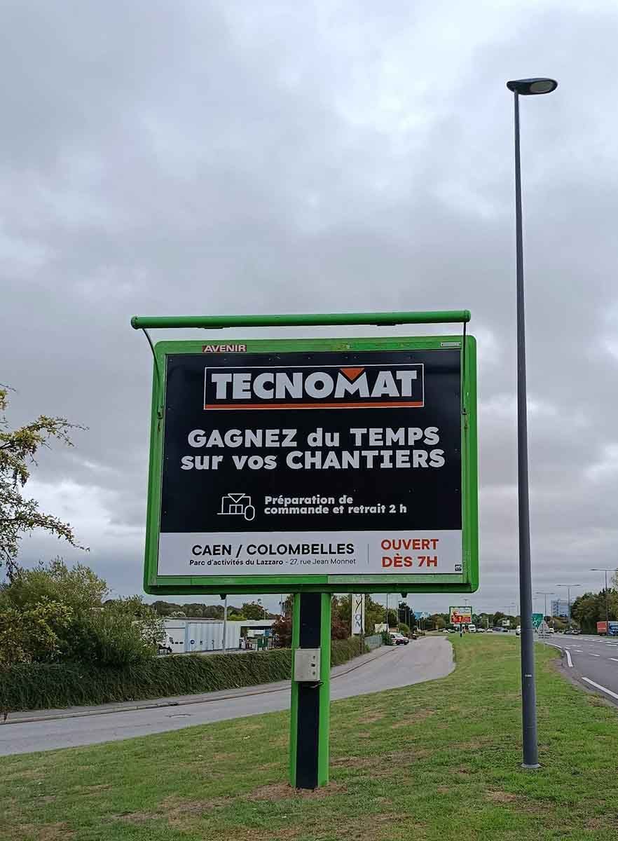 Panneau publicitaire pour TECNOMAT avec du texte en français, au bord d’une route, sous un ciel nuageux