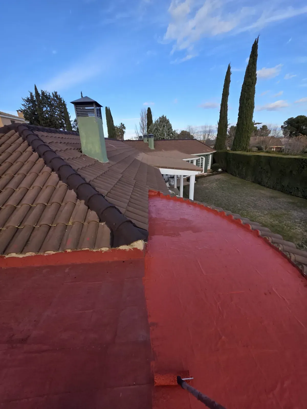 Un rodillo aplica sellador impermeabilizante rojo a una sección de techo plano contigua al tejado de una casa de tejas 