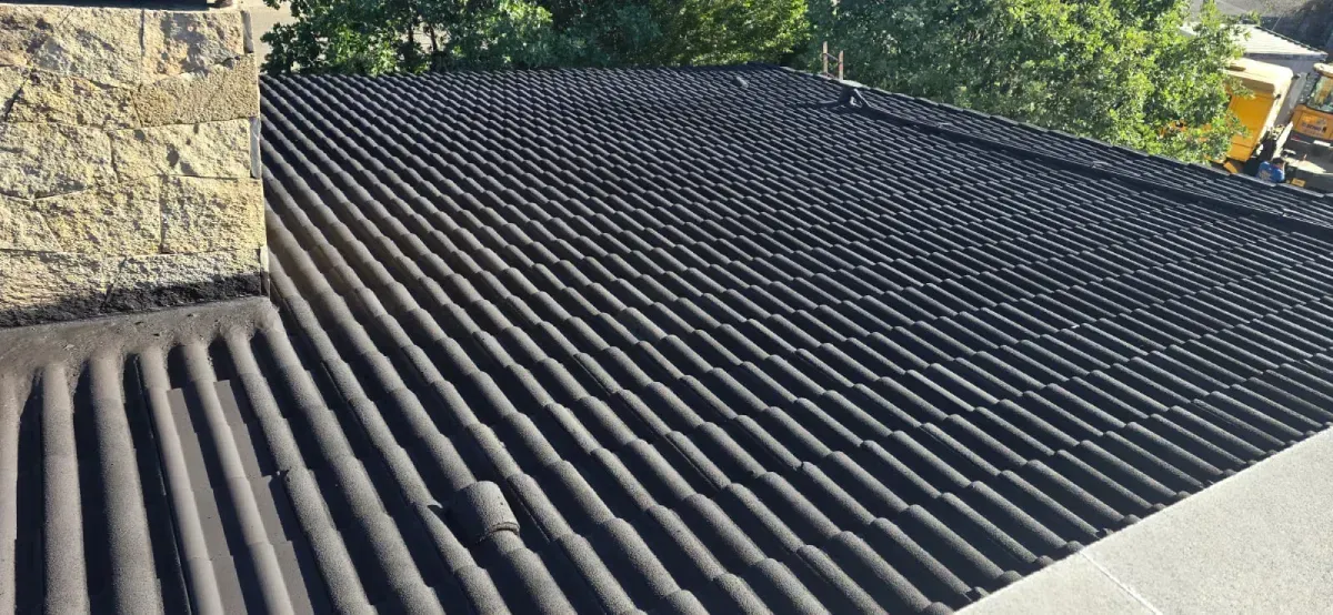 Vista desde un ángulo elevado de la superficie de un tejado de chapa ondulada oscura, junto a una chimenea de piedra.