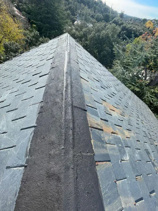 Un tejado inclinado con tejas de pizarra gris presenta una cumbrera de asfalto oscuro reparada