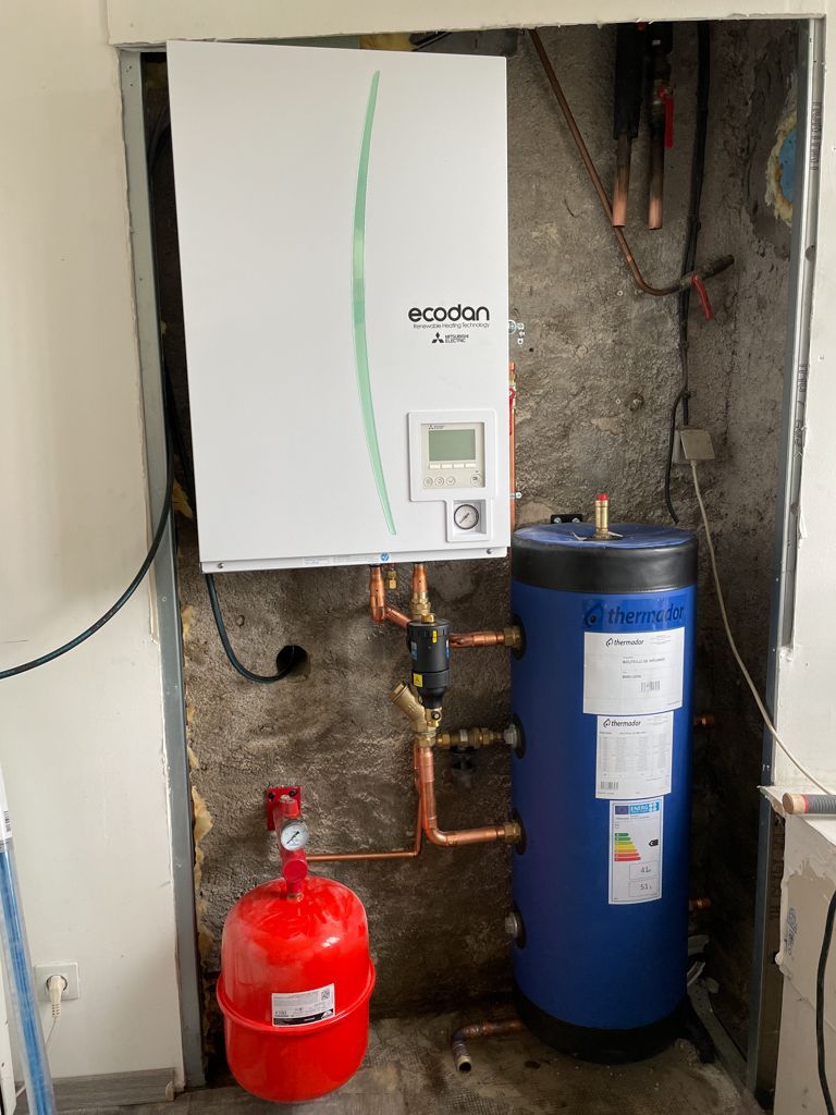 Une pompe à chaleur blanche et verte, des tuyaux en cuivre, un réservoir d'eau bleu et un vase d'expansion rouge à l'intérieur d'un mur encastré.
