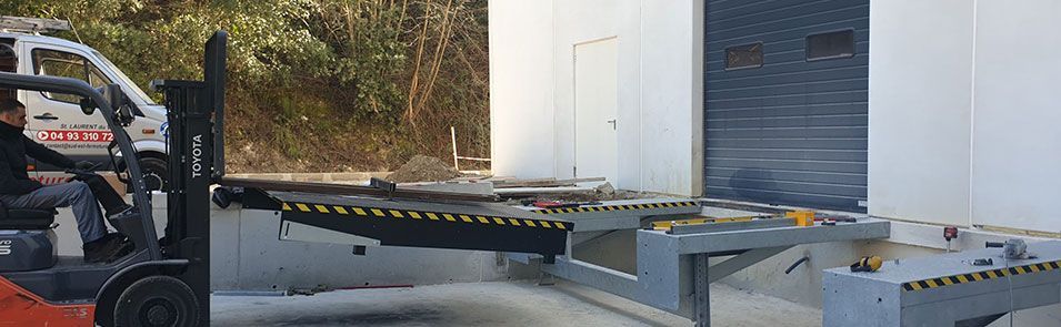 Déchargement par chariot élévateur sur un quai de chargement. Un opérateur manœuvre le chariot élévateur à côté d'un bâtiment blanc à porte grise.