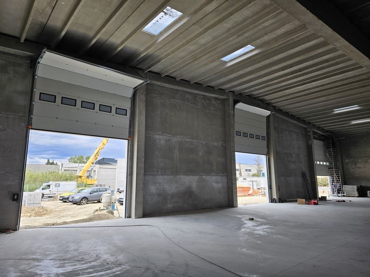 Intérieur d'un entrepôt avec portes sectionnelles ouvertes ; véhicules et engins de chantier visibles à l'extérieur.