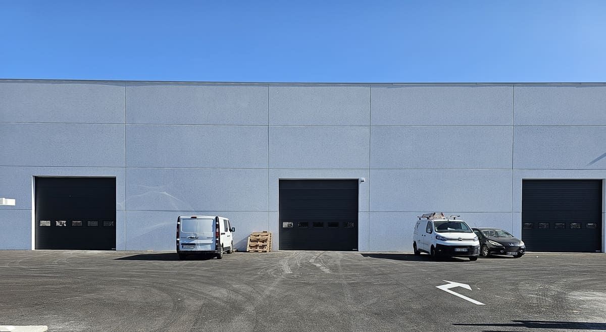 Extérieur d'un entrepôt avec trois portes de quai de chargement noires fermées ; des fourgonnettes blanches sont garées devant.