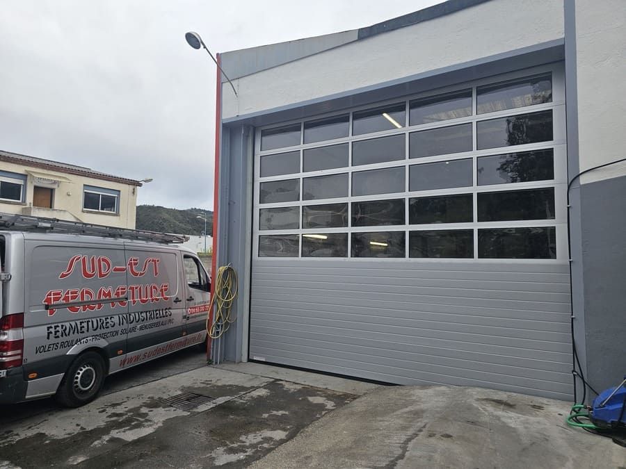 Porte de garage commerciale grise avec fenêtres, partiellement ouverte. Une camionnette est garée à gauche.