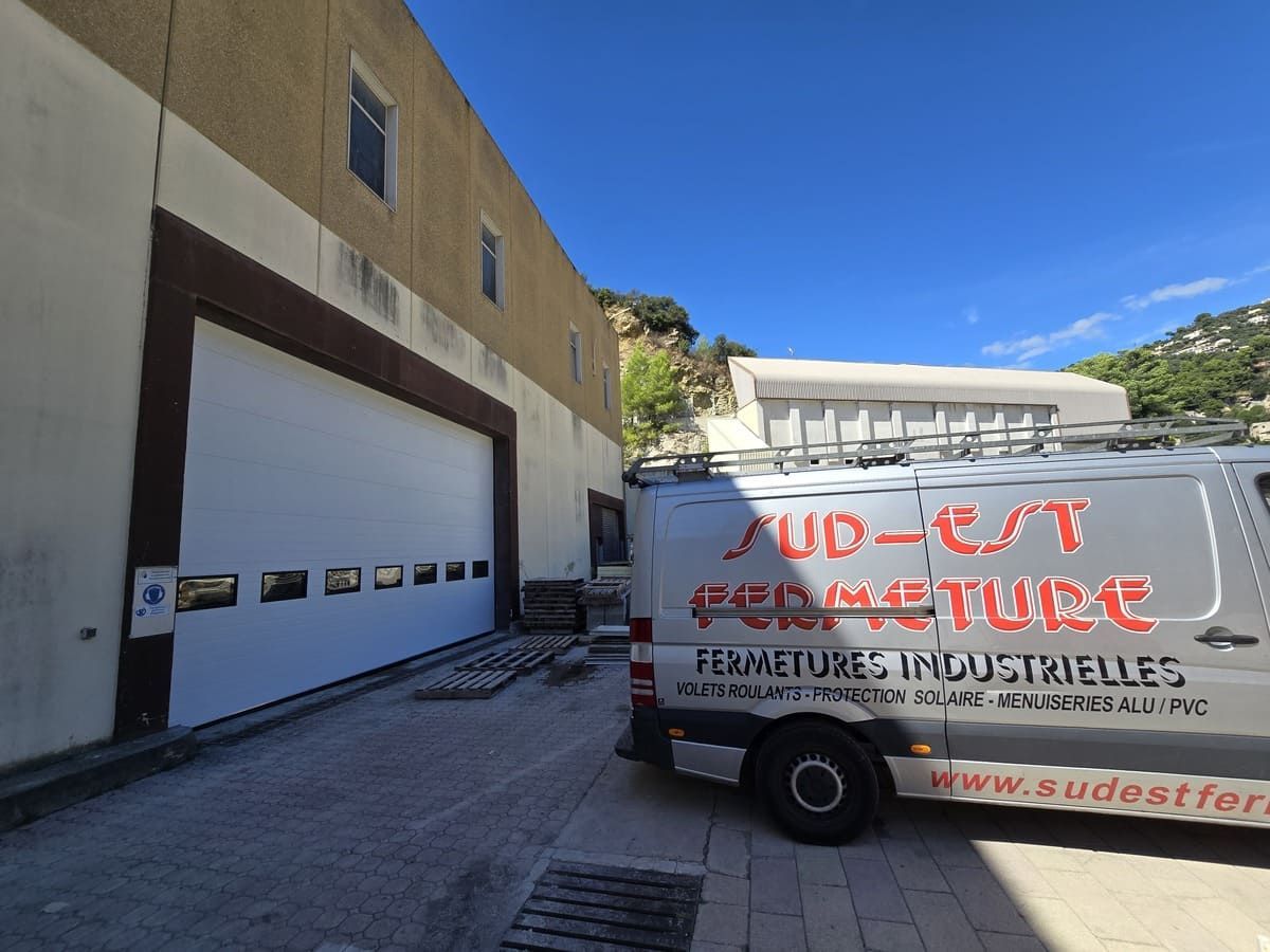 Porte industrielle blanche à côté d'un bâtiment beige ; camionnette « SUD-EST FERMETURE » garée devant.