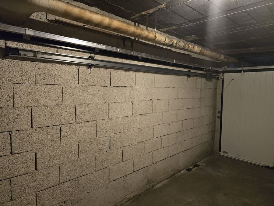 Mur en parpaings gris dans un garage, avec une porte de garage à droite. Le plafond est percé de tuyaux et d'un rail.
