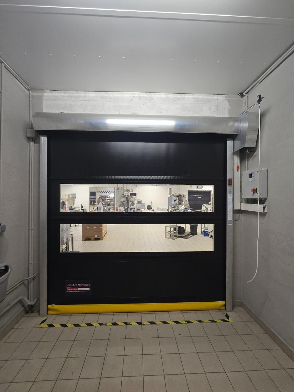 Porte sectionnelle industrielle noire avec fenêtres et bande de signalisation jaune dans une pièce aux murs blancs.