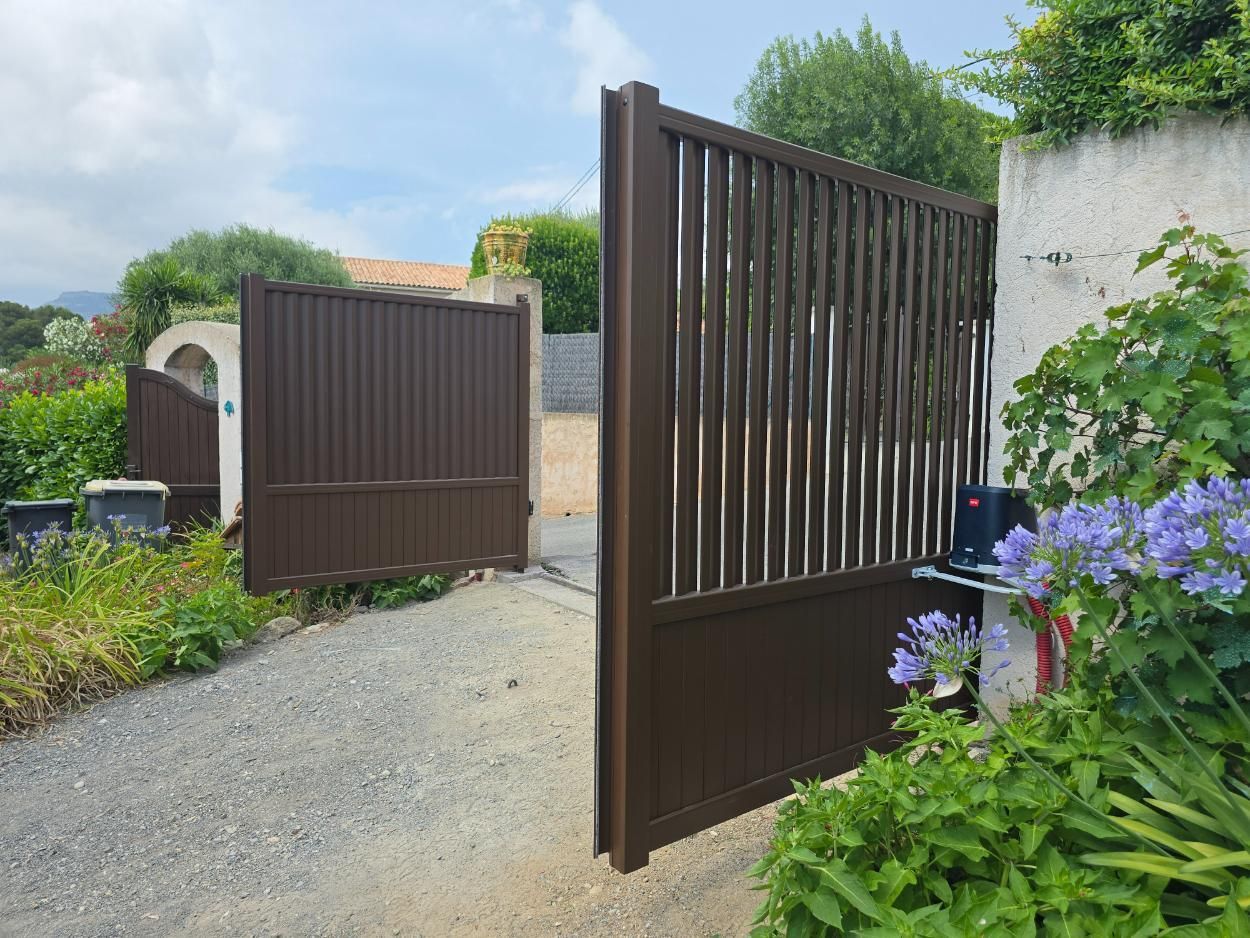 Un portail en métal brun, partiellement ouvert, laisse apparaître une allée de gravier. On aperçoit des plantes vertes et un bâtiment à l'arrière-plan.