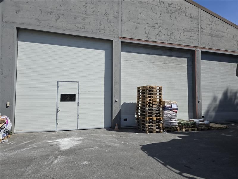 Vue extérieure d'un bâtiment industriel gris avec trois portes de garage. Des palettes en bois et des fournitures sont entreposées près d'une des portes.