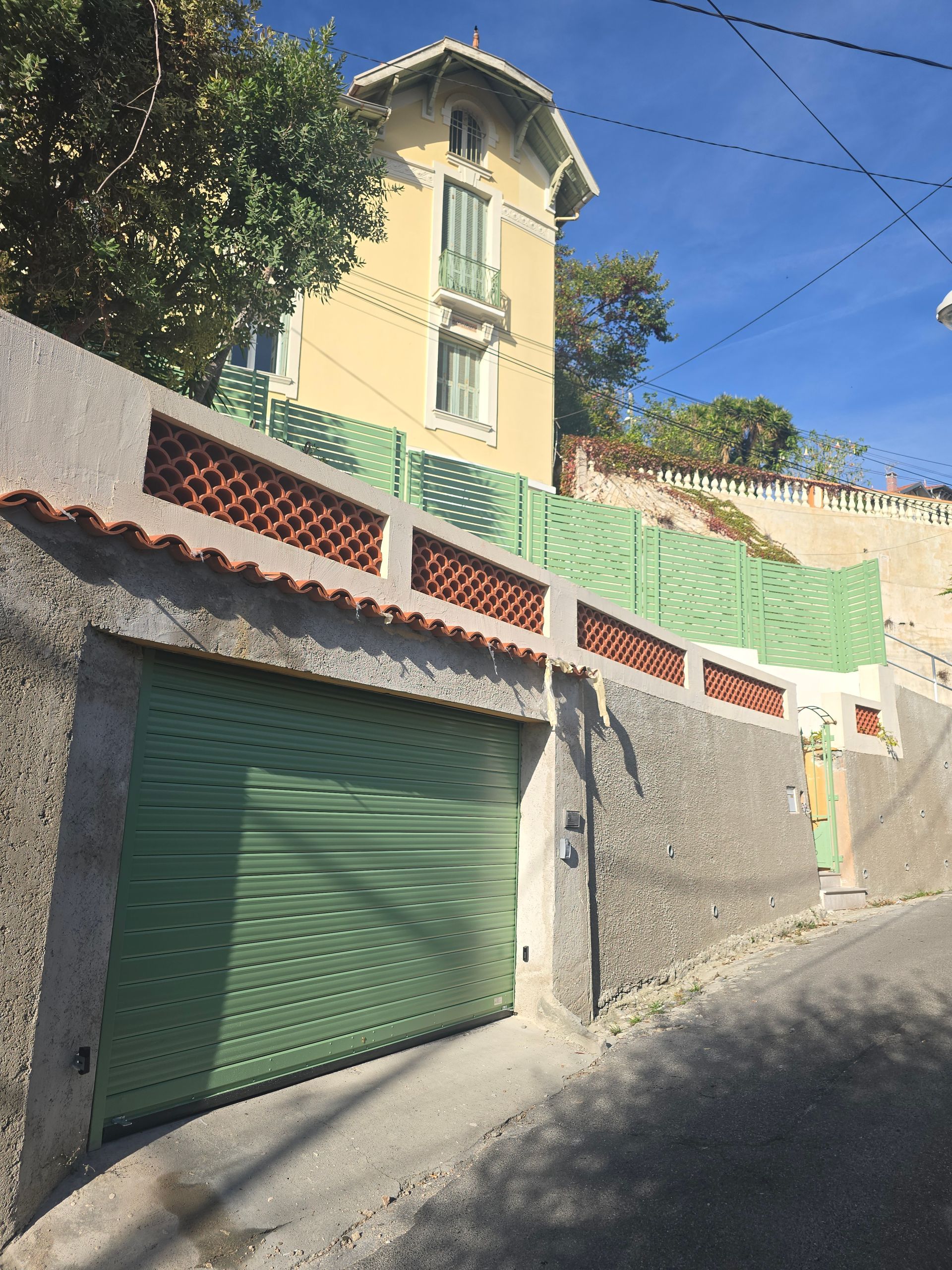 Porte de garage et mur verts, surmontés de panneaux verts et de tuiles rouges ; maison jaune à flanc de colline.