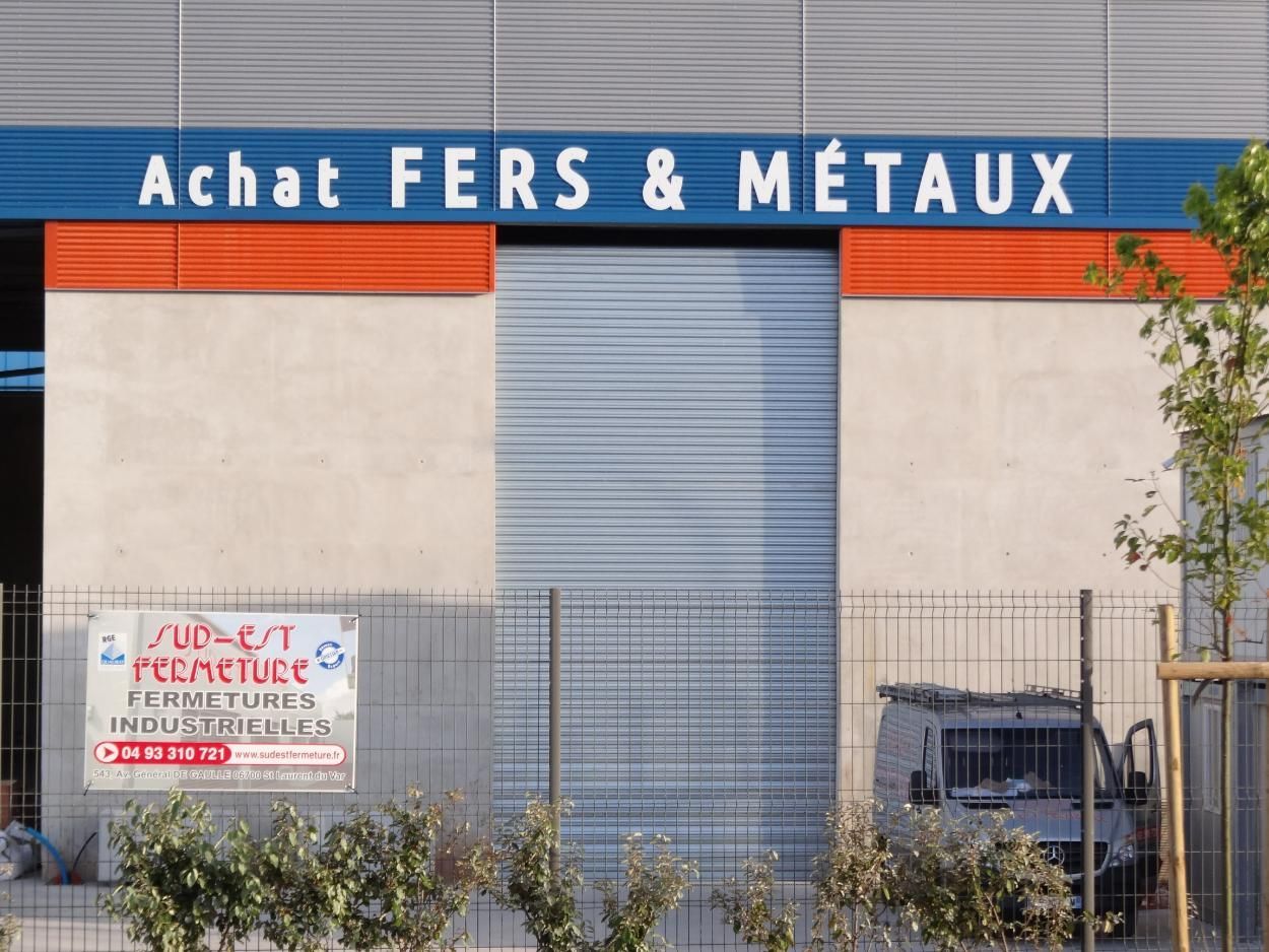 Façade du bâtiment avec des accents bleus et orange. Enseigne : « Achat FERS & MÉTAUX ». Volet roulant fermé.