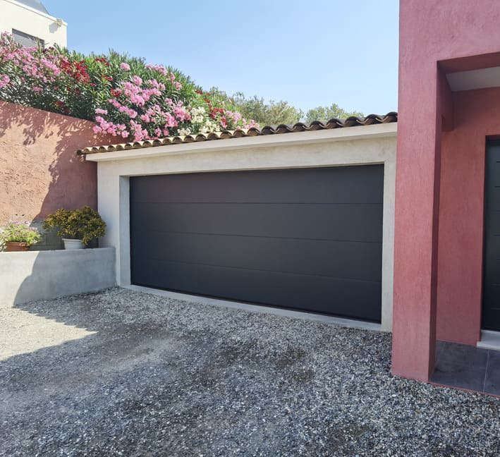 Porte de garage grise, allée de gravier et façade rouge du bâtiment par une journée ensoleillée.