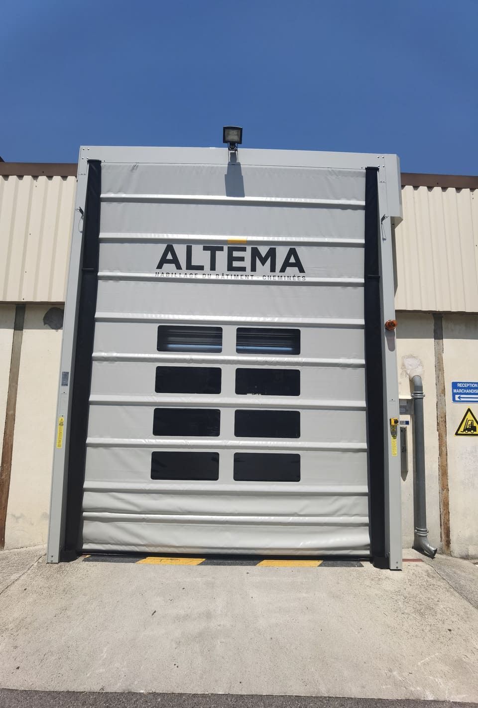 Porte industrielle grise avec inscription « ALTEMA » et plusieurs fenêtres noires, sur un quai de chargement en béton.