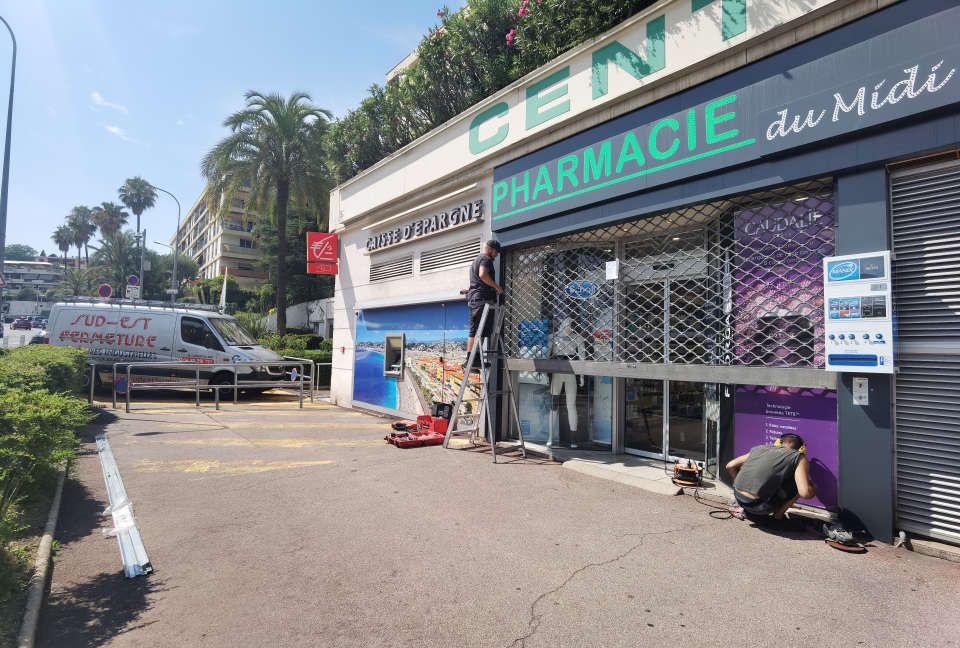 Des ouvriers installent des panneaux de signalisation à l'extérieur d'une pharmacie équipée d'un rideau métallique de sécurité ; une camionnette blanche est garée à proximité.