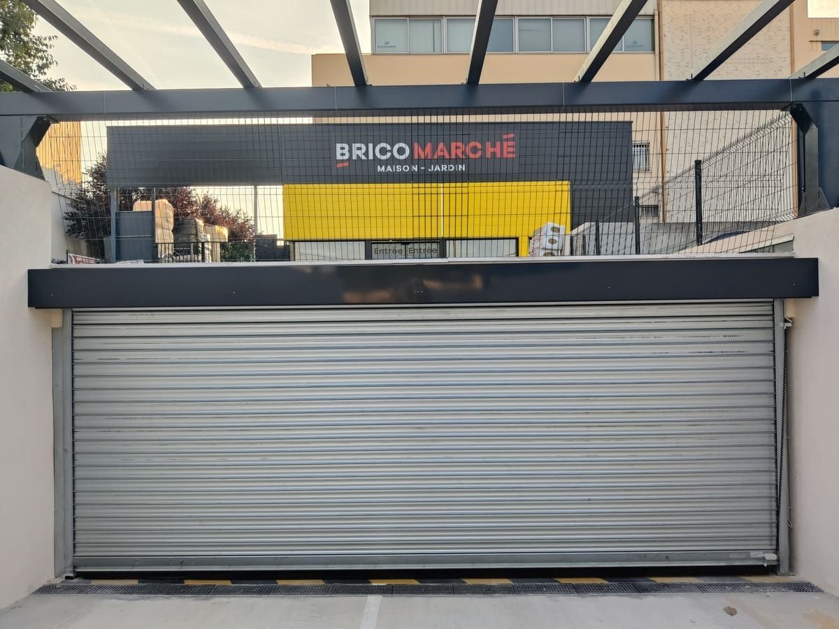 Volet métallique fermé devant un magasin Brico Marché ; façade du bâtiment noire et jaune.