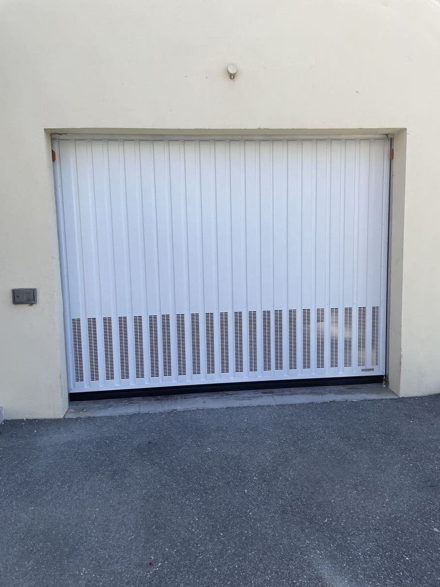 Porte de garage accordéon blanche, partiellement ouverte sur un bâtiment beige clair. Asphalte en dessous.
