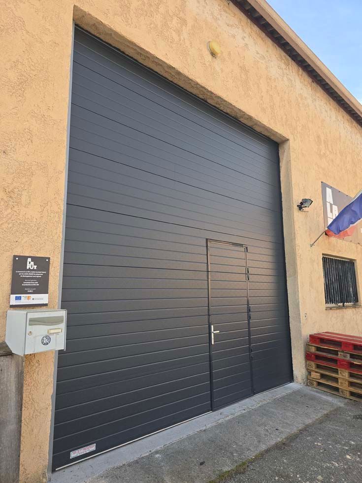 Grande porte d'entrepôt gris foncé avec une porte plus petite encastrée. Extérieur d'un bâtiment beige.