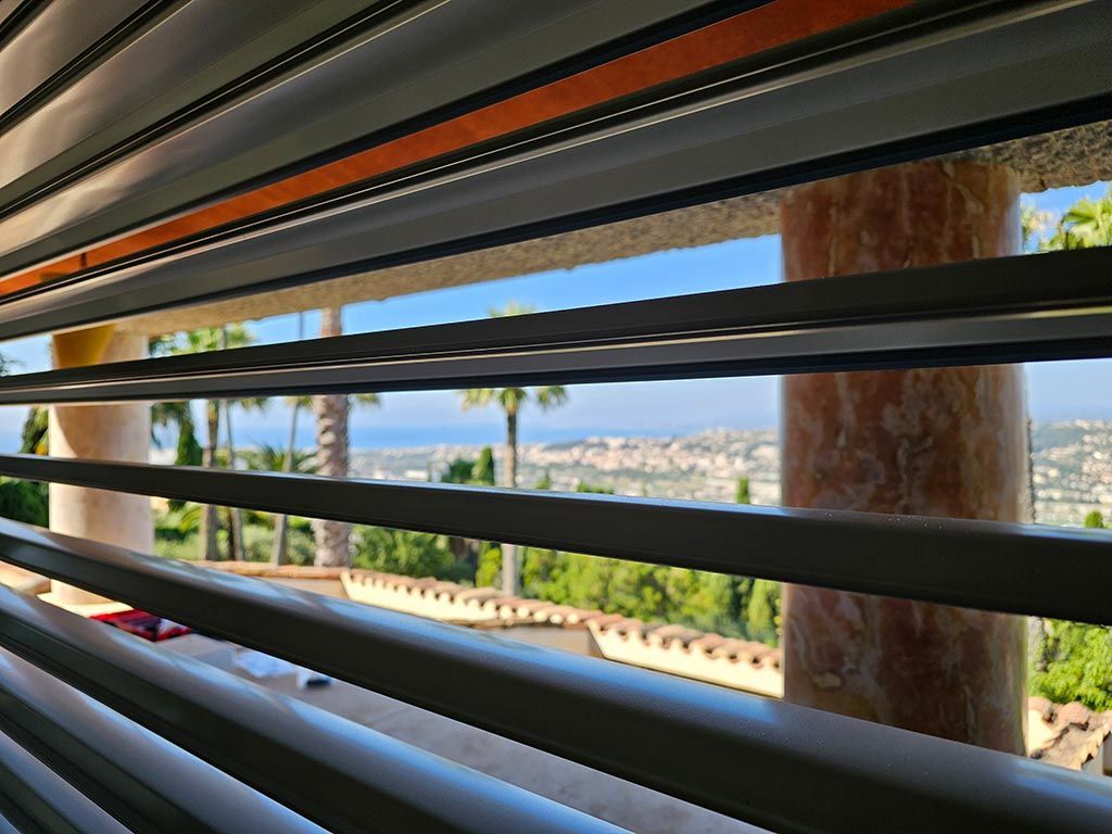 Vue à travers les stores partiellement ouverts : paysage urbain, ciel bleu, palmiers et pilier de marbre.