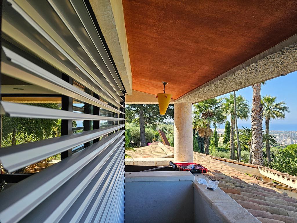 Vue depuis un balcon aux stores blancs à lamelles, donnant sur des palmiers et un paysage urbain au loin.