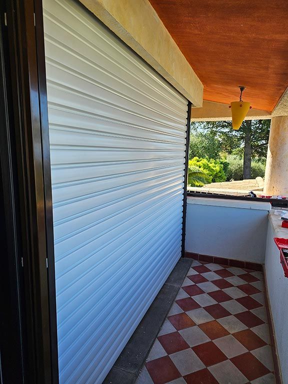 Volet roulant blanc fermé sur une terrasse extérieure avec un sol en carrelage à damier et un luminaire jaune.