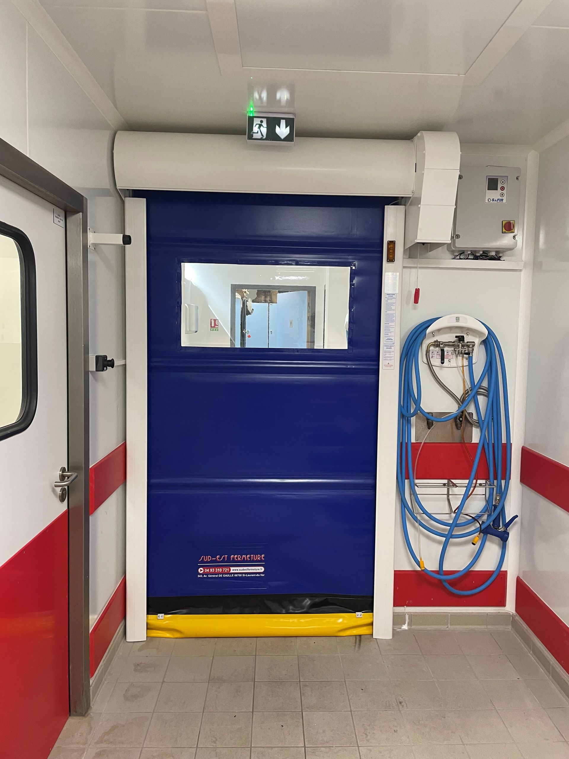 Porte sectionnelle industrielle bleue et jaune dans une pièce blanche avec une petite fenêtre, panneau de sortie au-dessus.