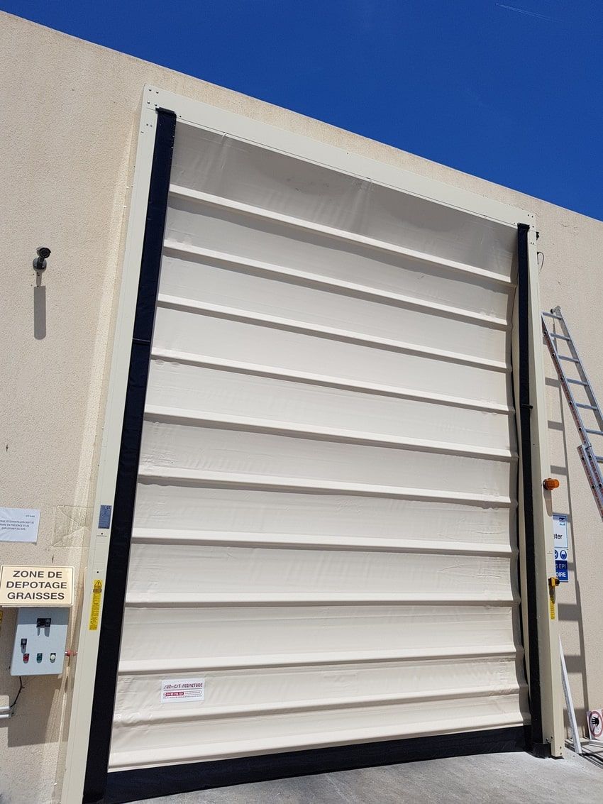 Porte sectionnelle beige sur un bâtiment beige avec joints latéraux noirs. Une échelle et un boîtier électrique sont visibles.