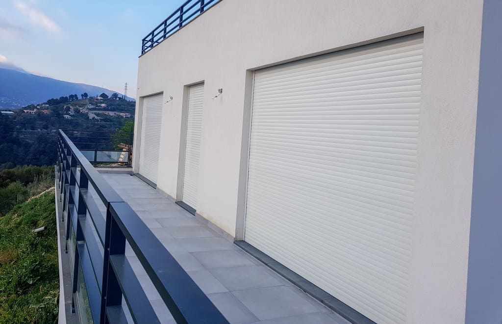 Bâtiment aux volets blancs fermés et à la balustrade donnant sur un paysage montagneux.