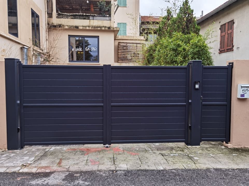 Portail à lattes horizontales gris foncé, installé entre des piliers, sur un trottoir en briques. Bâtiment visible derrière le portail.