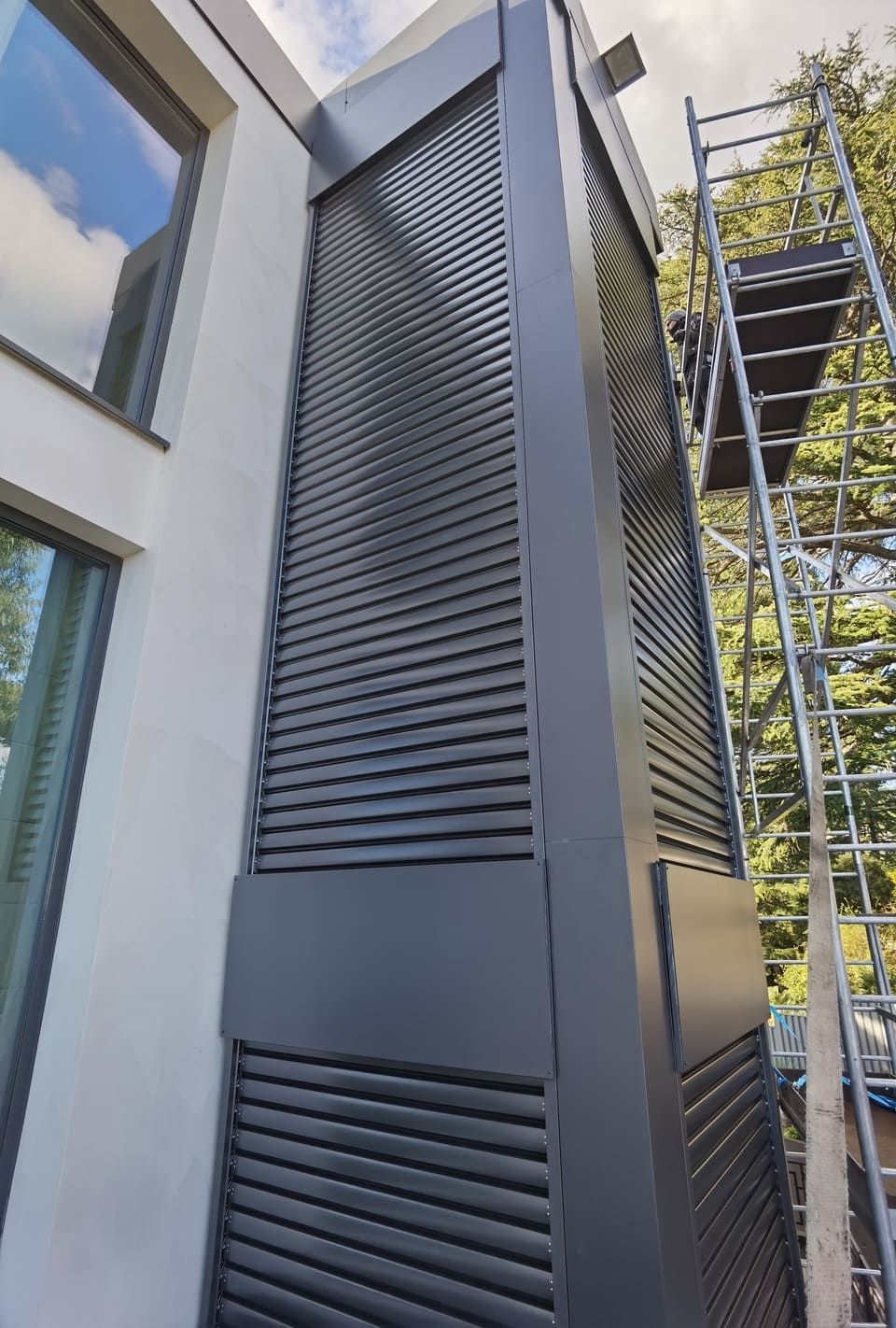 Structure grise à persiennes adossée à un bâtiment blanc ; échafaudages à proximité.