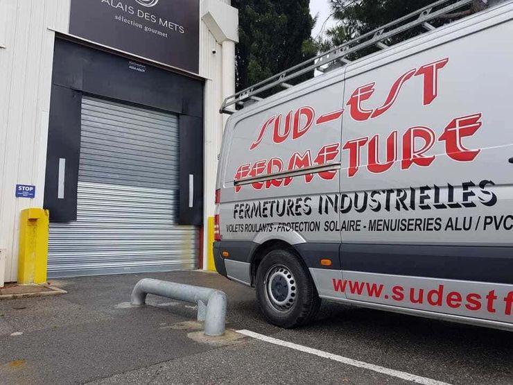 Une camionnette argentée portant le logo « Sud-Est Fermeture » est garée près d'un bâtiment dont le rideau métallique de sécurité argenté est fermé.