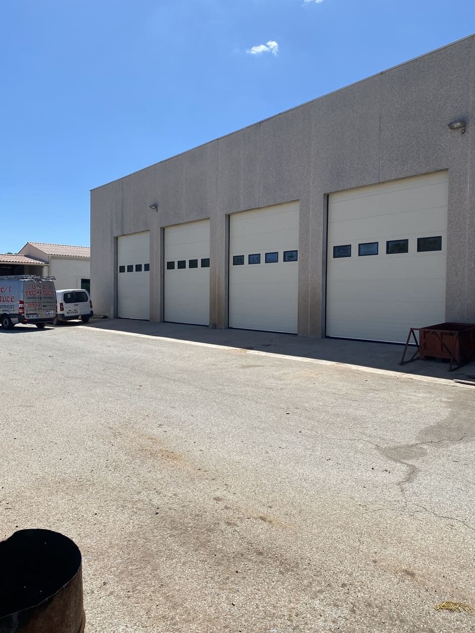 Extérieur d'un bâtiment beige avec quatre portes de garage, un terrain gravillonné devant et un ciel bleu.