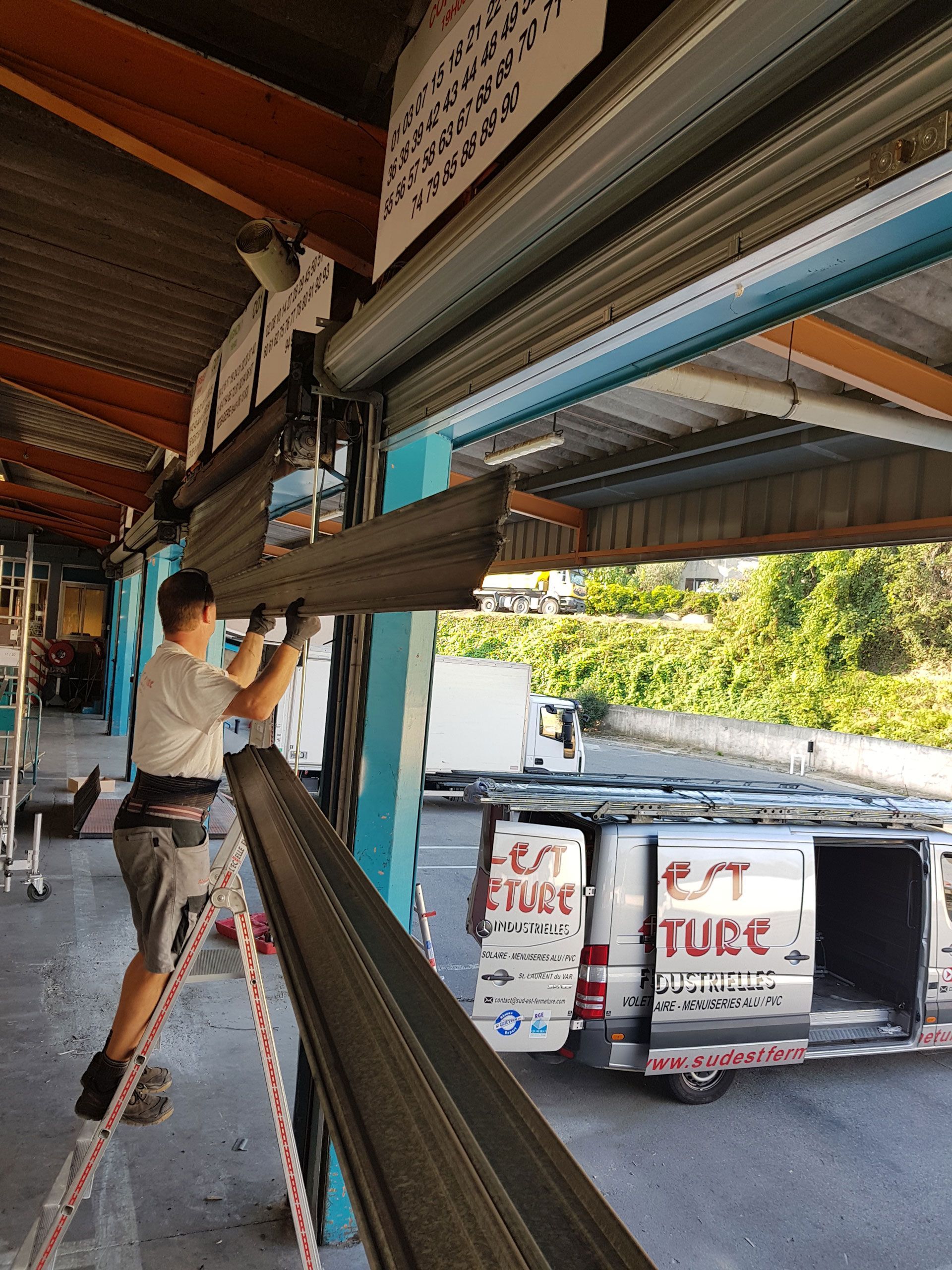 Un homme sur une échelle répare un rideau métallique gris endommagé ; une camionnette blanche portant le logo « EST TURC » est garée à proximité.