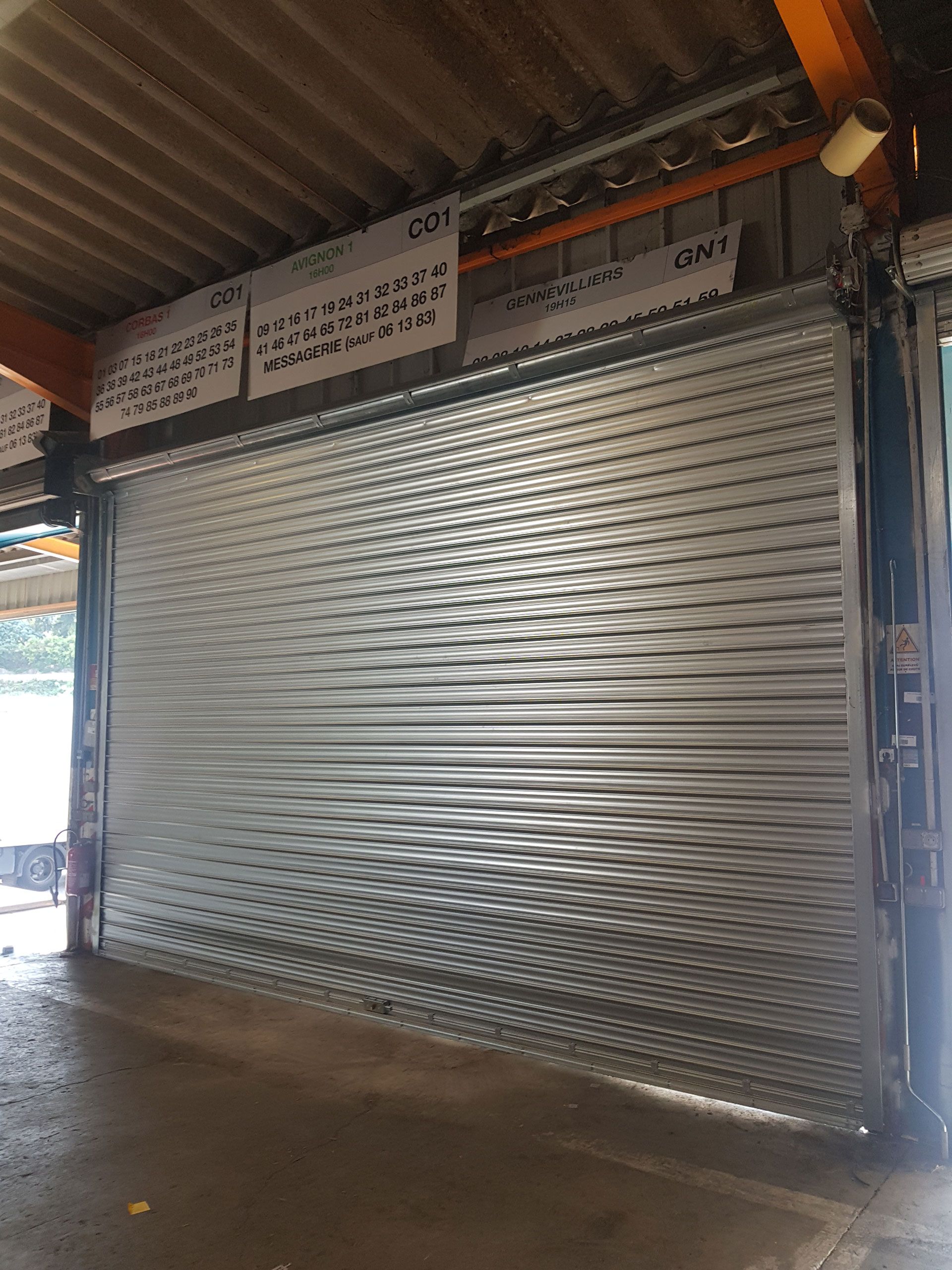 Porte sectionnelle en métal argenté, partiellement fermée. Sous un toit en tôle ondulée, vue intérieure.