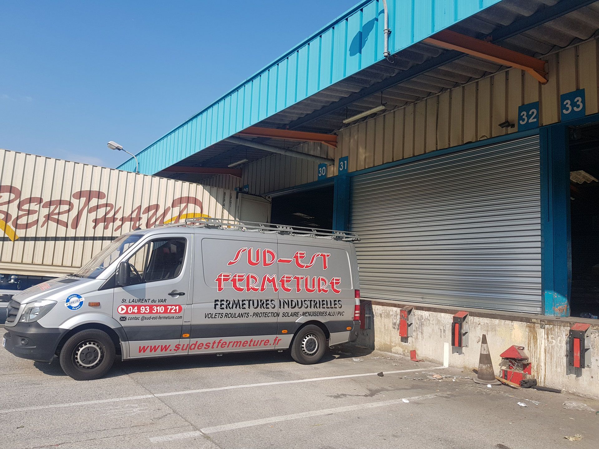 Camionnette stationnée au quai de chargement. Le bâtiment possède un auvent bleu et une grande porte. On peut lire sur la camionnette : « SUD-EST FERMETURE ».