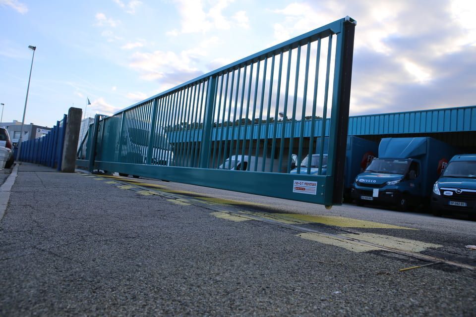 Un portail coulissant vert bloque l'accès à l'entrée de l'entreprise, des camionnettes bleues sont garées à proximité.