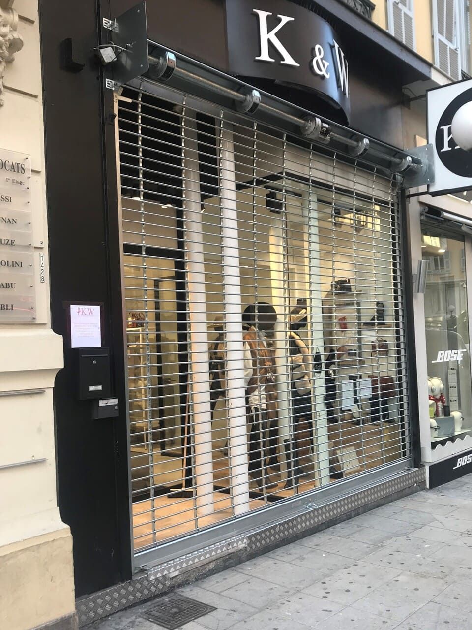 Une devanture de magasin avec un rideau métallique de sécurité baissé, laissant apparaître les articles à l'intérieur. Enseigne « K & W » au-dessus.
