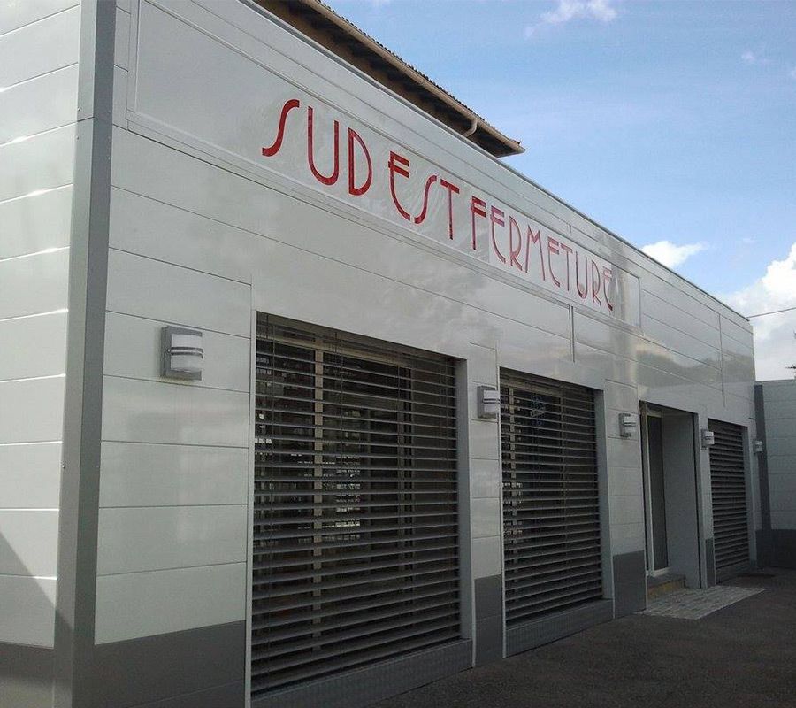 Bâtiment blanc avec l'inscription « SUD EST FERMETURES » en rouge, portails de sécurité fermés.