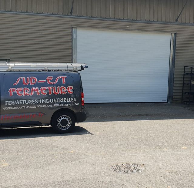 Une porte de garage blanche jouxte un bâtiment gris. Une camionnette est garée à gauche ; on peut lire « Sud-Est Fermeture » sur le côté.